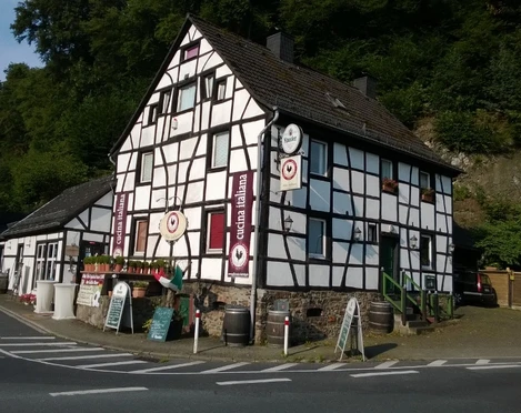 Fachwerkhaus mit Gaststätte, von Wald umgeben, bei strahlendem Sonnenschein an einer Straßenecke.