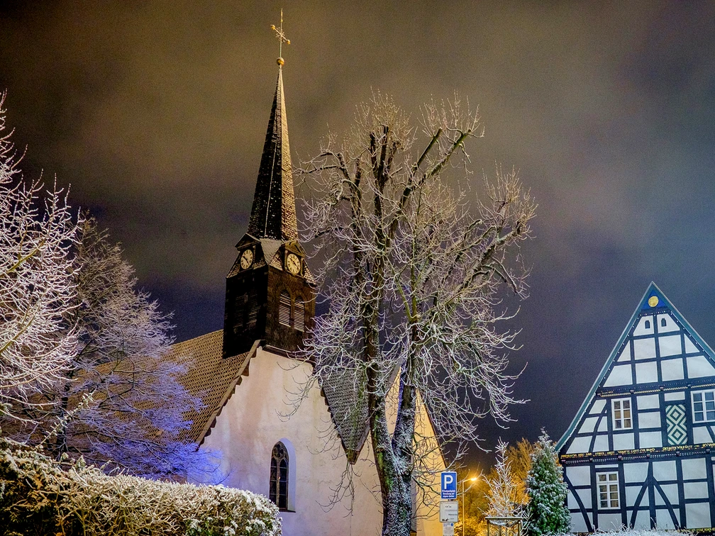Die winterliche Kirche St. Stephan in verschneiter, abendlicher Landschaft, flankiert vom Fachwerkhaus.