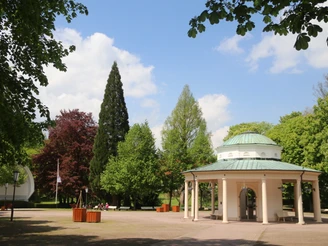 Brunnentempel mit Musikmuschel Brunnentempel mit Musikmuschel inmitten eines gepflegten Parks mit grünen Bäumen und blauen Himmel.