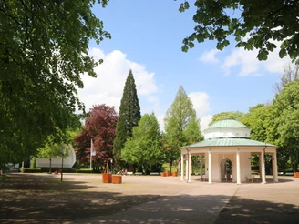 Brunnentempel mit Musikmuschel Ein historischer Pavillon mit grüner Kuppel steht in einem parkähnlichen Umfeld voller Bäume.