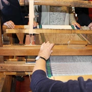 Webstuhl Ein traditioneller Webstuhl mit Holzstruktur, umgeben von Menschen, die das Weben beobachten oder helfen.