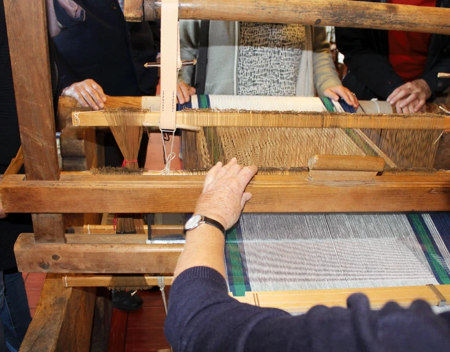 Webstuhl Ein traditioneller Webstuhl mit Holzstruktur, umgeben von Menschen, die das Weben beobachten oder helfen.