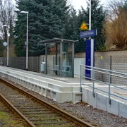Ein kleiner Bahnhof mit modernem Wartebereich, Geländer, Bäumen und einem Weg entlang der Gleise.