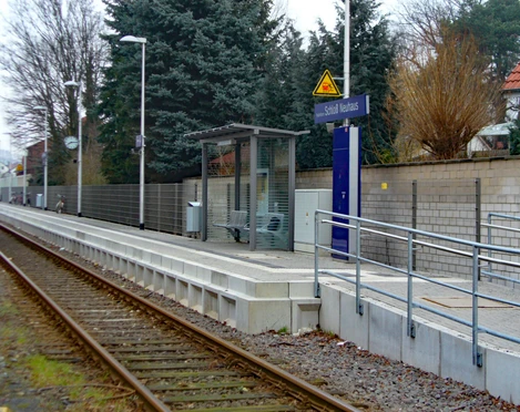 Ein kleiner Bahnhof mit modernem Wartebereich, Geländer, Bäumen und einem Weg entlang der Gleise.