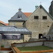 Steinbau mit Satteldach und quadratischem Turm, Flachdachgebäude und Bannerschrift im Vordergrund.