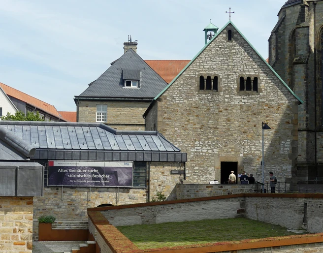 Steinbau mit Satteldach und quadratischem Turm, Flachdachgebäude und Bannerschrift im Vordergrund.