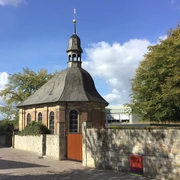 Alexiuskapelle in Paderborn: Backsteingebäude mit rundem Dach und Turm, umgeben von Bäumen im Sonnenschein.