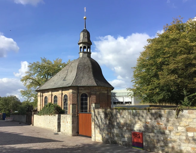 Alexiuskapelle in Paderborn: Backsteingebäude mit rundem Dach und Turm, umgeben von Bäumen im Sonnenschein.