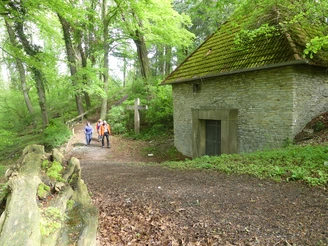 Zwei Wanderer nahe einem historischen, von Bäumen umgebenen Mausoleum mit einem großen Holzkreuz.