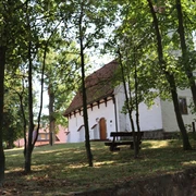 Eine weiße Kapelle mit rotem Ziegeldach, umgeben von Bäumen, eine Holzbank im Vordergrund.