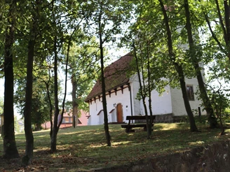 Eine weiße Kapelle mit rotem Ziegeldach, umgeben von Bäumen, eine Holzbank im Vordergrund.