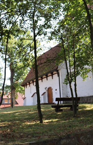 Eine weiße Kapelle mit rotem Ziegeldach, umgeben von Bäumen, eine Holzbank im Vordergrund.