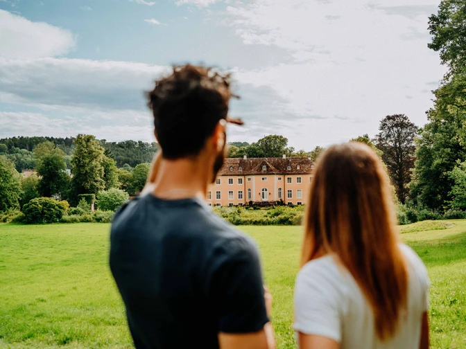 Ein Mann und eine Frau blicken auf Schloss Rheder, umgeben von üppigen grünen Wiesen und Wäldern.