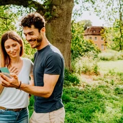 Teutoburger Wald-Brakel-Schloss Rheder  Zwei Menschen stehen lächelnd auf einer Wiese und betrachten ein Smartphone, ein Schloss ist im Hintergrund.