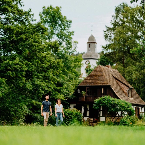 Teutoburger Wald-Brakel-Schloss Rheder Zwei Personen spazieren vor einem historischen Fachwerkhaus mit Turm, umgeben von üppigem Grün.