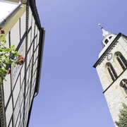 Giebel des Historischen Rathauses Wiedenbrück Fachwerkhaus, rote Blüten und Kirche mit Uhrturm, blauer Himmel im Hintergrund.