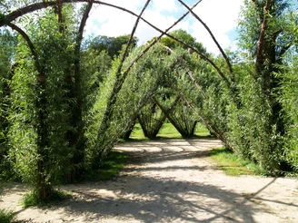 Weidenpalais Rheder Ein kunstvoll gewölbtes Bauwerk aus geflochtenen Weiden, umgeben von üppigem Grün im offenen Freiraum.