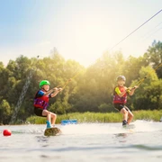Zwei Kinder auf Wakeboards, gezogen von einem Seil über die Wasseroberfläche, unter strahlendem Sonnenschein.