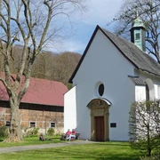 Kleine, weiße Kapelle neben einer großen Scheune, mehrere Personen sitzen im Grünen vor der Kapelle.