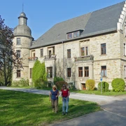 Eine Gruppe von Menschen spaziert vor einer historischen Schlossgebäude mit Turm und Gartenanlage.