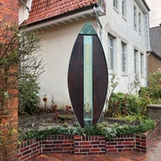 Skulptur Das Portal Bücken Eine hohe, moderne Skulptur in Form eines tropfenförmigen Ovals neben einem roten Backsteingebäude.