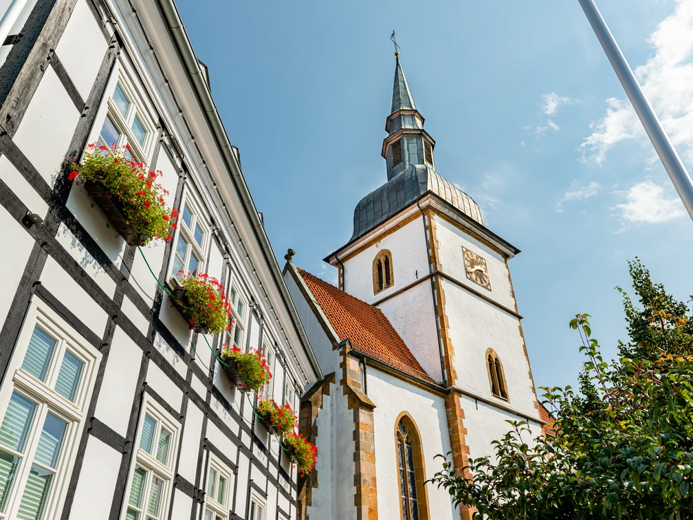 Historisches Fachwerkgebäude neben der St. Ursula-Kirche mit blühenden Pflanzen im Vordergrund.