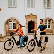 Teutoburger Wald-Brakel-Tourist Info ┬® Teutoburger Wald Tourismus (5).jpg Zwei Personen mit E-Bikes stehen vor einem historischen Gebäude mit Fachwerkelementen.