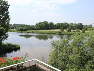 Hotel Weserschlößchen Nienburg Flusslandschaft mit grünen Ufern und Bäumen, vereinzelt roten Blumen im Vordergrund, leichte Bewölkung.