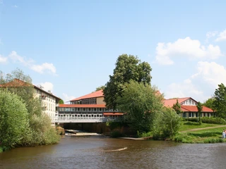 Hotel-Weserschlößchen-Nienburg Hotel mit rotem Dach an Flussufer, umgeben von grünen Bäumen, blauer Himmel im Hintergrund.
