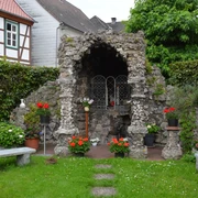Eine mit Blumen geschmückte Grotte aus Naturstein, umgeben von gepflegtem Grün und zwei Steinbänken.