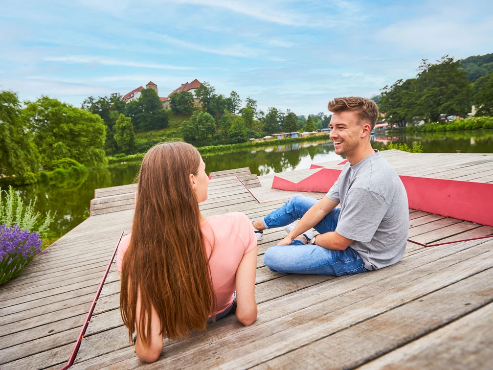 Relaxen am Charlottensee in Bad Iburg Ein Mann und eine Frau sitzen entspannt auf einem Holzsteg am See, im Hintergrund grüne Bäume.