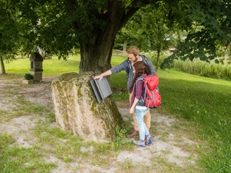 Eisernes Buch Andepen Zwei Menschen betrachten ein offenes, metallisches Buch auf einem Moos-bedeckten Stein unter einem Baum.