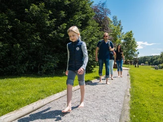 Erlebnis-Barfußpfad in Bad Wünnenberg Menschen spazieren barfuß auf einem Kiesweg durch einen grünen Park bei klarem Himmel und Sonnenschein.