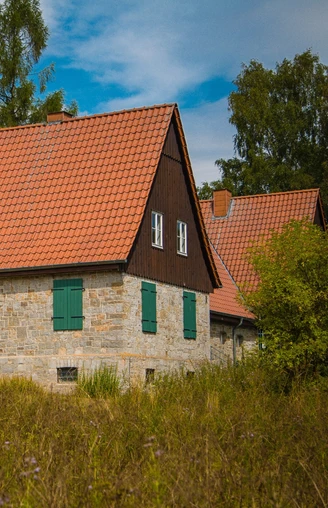 Aahäuser bei Bleiwäsche Ein Steinhaus mit rotem Ziegeldach und grünen Fensterläden, umgeben von Bäumen unter blauem Himmel.