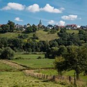 Grüne Wiesen mit einem Pfad führen zu einem Dorf auf einem Hügel, umgeben von Bäumen.