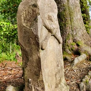 Steinerner Totenschädel-Monolith im Wald, umrahmt von Bäumen und Wurzeln an einem sonnigen Tag.