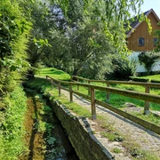 Mühlengraben in Brakel-Bellersen Kleiner Bachlauf mit Holzgeländer, angrenzenden Wiesen und Bäumen, im Hintergrund ein Fachwerkhaus.