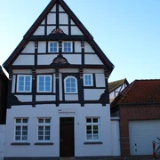 Fachwerkhaus mit weißer Fassade und dunklem Holzgebälk, umgeben von Ziegelbauten, vor blauem Himmel.