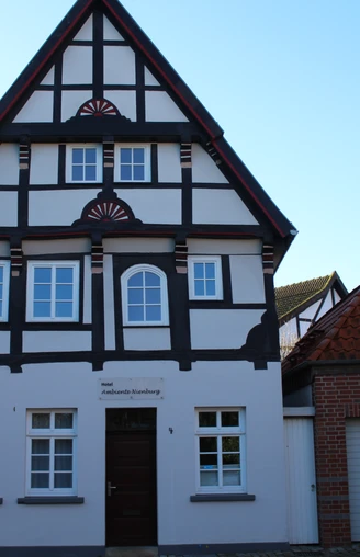 Fachwerkhaus mit weißer Fassade und dunklem Holzgebälk, umgeben von Ziegelbauten, vor blauem Himmel.
