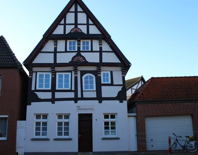 Fachwerkhaus mit weißer Fassade und dunklem Holzgebälk, umgeben von Ziegelbauten, vor blauem Himmel.
