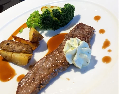 Fleischgericht Ein Stück gegrilltes Rindersteak mit Kräuterbutter, Kartoffelecken und Brokkoli auf weißem Teller.