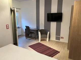 Modern eingerichtetes Hotelzimmer mit einem Schreibtisch, Stuhl, Sessel und einem Flachbildfernseher.