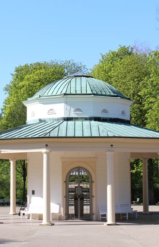 Brunnentempel im Historischen Kurpark Runder Pavillon mit Säulen und grüner Kuppel in einem grünen Kurpark, umgeben von Bäumen.