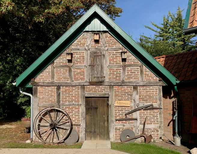 image00015.jpeg Altes Fachwerkhaus mit Holzreifen und Pflug an der Fassade unter blauem Himmel.