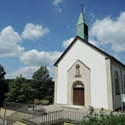 St. Josef Kapelle in Feldrom mit weißer Fassade, grünem Turm und umgeben von üppigem Grün.