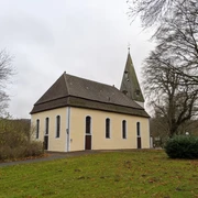 Kirche Stapelage_2©Tourist-Information Lage.jpg Die Kirche Stapelage mit spitzem Turm, umgeben von Bäumen und Rasen, unter bedecktem Himmel.