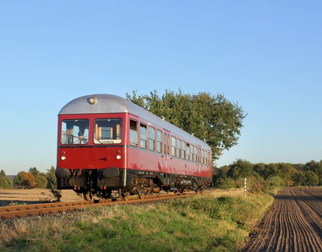 Bleckeder-kleinbahn-triebwagen-foto_ccbysa_foto©Bleckeder-kleinbahn.jpg Ein roter historischer Triebwagen der Bleckeder Kleinbahn fährt auf Gleisen durch eine ländliche Herbstlandschaft.