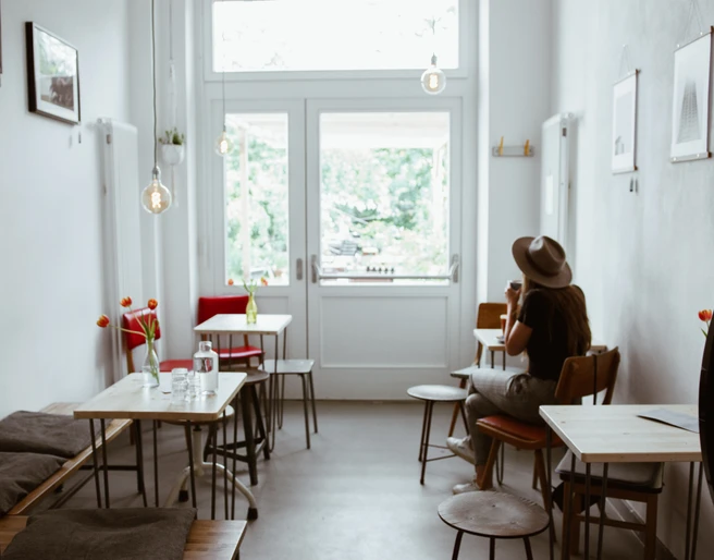 Innenansicht Café Analog - Cafés in Leipzig Zusehen sind die Sitzplätze des Cafés: Kleine Tische und Bänke aus Holz sowie kleine Bilder, die an der Wand hängen. Der Raum wird von einem großen Glasfenster erhellt, Cafe, Gastronomie, Freizeit
