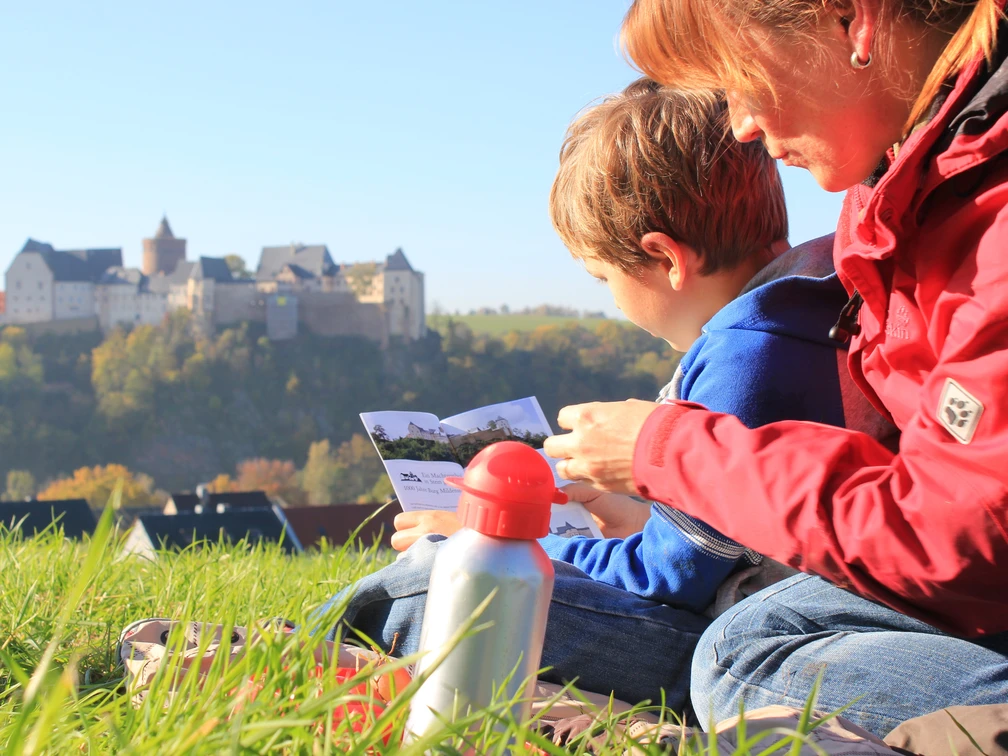 Burg Mildenstein - Sehenswürdigkeiten in der Leipzig Region Eine Frau mit Kind sitzt mit einer Broschüre in der Hand auf einer Wiese, im Hintergrund erhebt sich Burg Mildenstein, Auflug, Familie,Paní s dítětem sedí na louce s brožurou v ruce, v pozadí se tyčí hrad Mildenstein, výlet, rodina,A woman and her child sit on the grass looking at a brochure with Mildenstein Castle in the background at the top of the hill, excursion, family,Une femme et son enfant assis dans l’herbe, une brochure à la main, avec en arrière-plan le château de Mildenstein, excursion, famille,Kobieta z dzieckiem siedzą na łące, w ręku trzymają broszurę turystyczną, w tle rysuje się zamek Mildenstein, wycieczka, rodzina