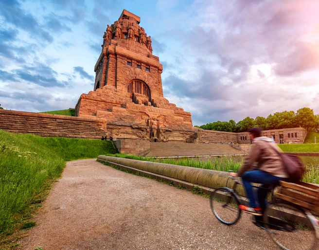 Völkerschlachtdenkmal - Sehenswürdigkeiten in Leipzig Blick auf das von der warmen Sonne angestrahlte Völkerschlachtdenkmal Leipzig, während ein Fahrradfahrer am See der Tränen vorbeiradelt, eine wahre Sehenswürdigkeit und ein besonderes Stück der Architektur in Leipzig, Ausflug, Sehenswürdigkeiten, Kultur